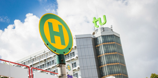 Bushaltestellenschild mit Mensabrücke und Mathetower im Hintergrund.